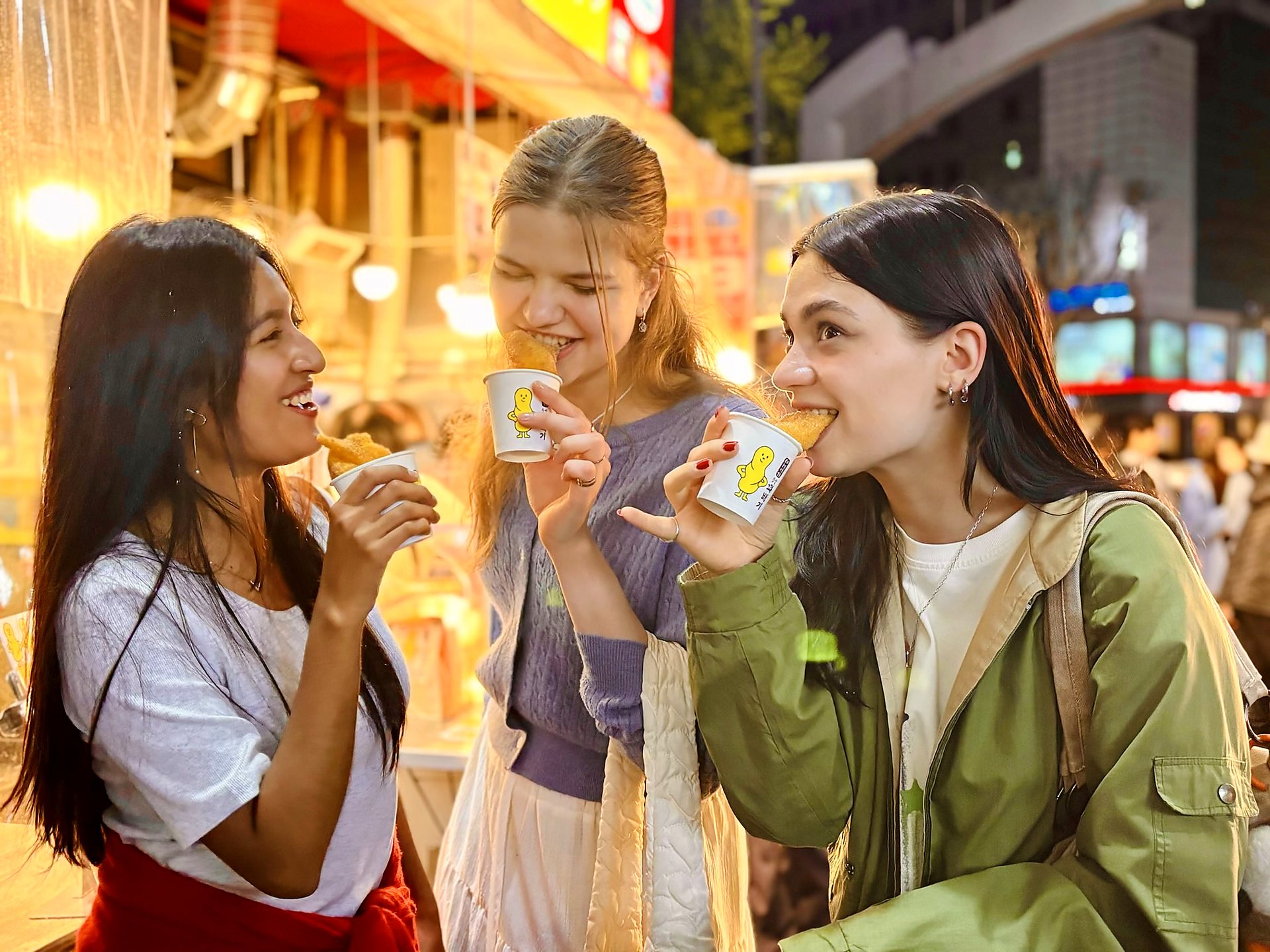 Three guests eating street food snacks at a lit Gwangjang Market stall at night