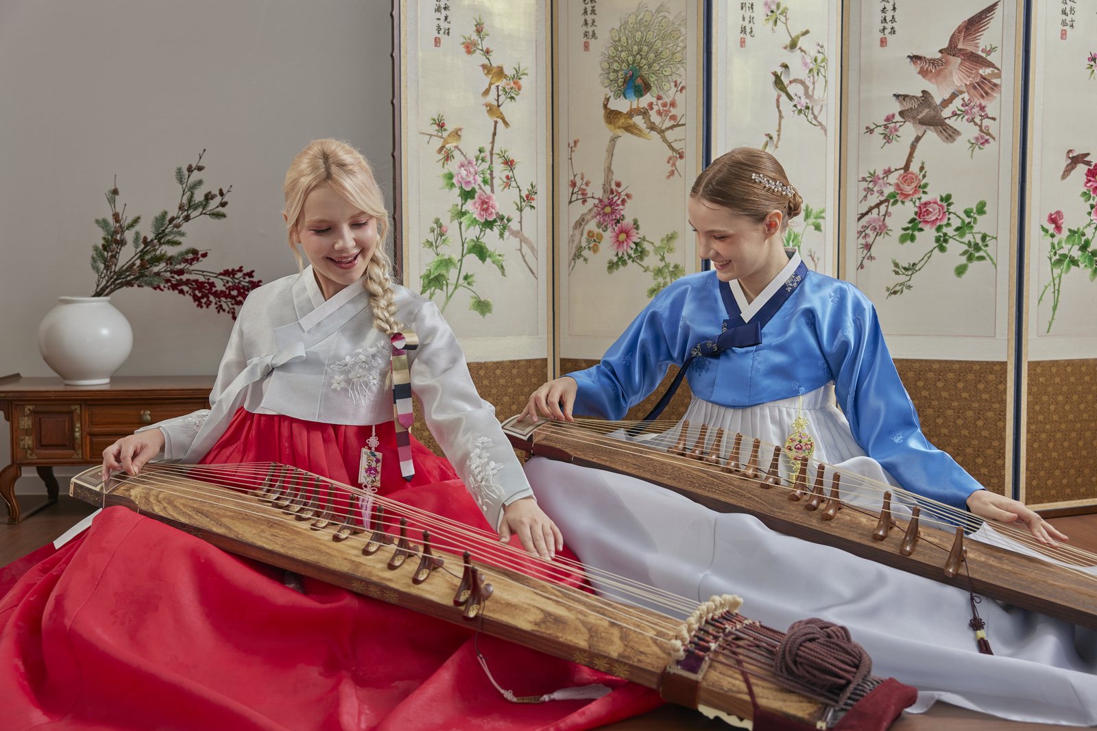 Two guests in hanbok seated playing gayageum in a studio with painted folding screens